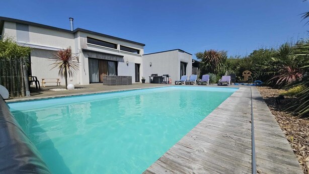 Un espace piscine moderne avec une terrasse en bois, des chaises longues et un bâtiment doté de grandes fenêtres et d’un patio couvert.