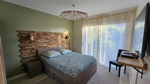 Une chambre avec une tête de lit en pierre, un lit avec une literie à motifs, un bureau avec un ordinateur et une grande fenêtre avec des rideaux.