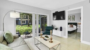 A modern living room with a sofa, a coffee table, and a TV. There is a door leading to a bedroom with a bed and a TV. - 340A - Boca Retreat w/ Pool, Parking, Near Evert Tennis & Shops (Boca Raton)