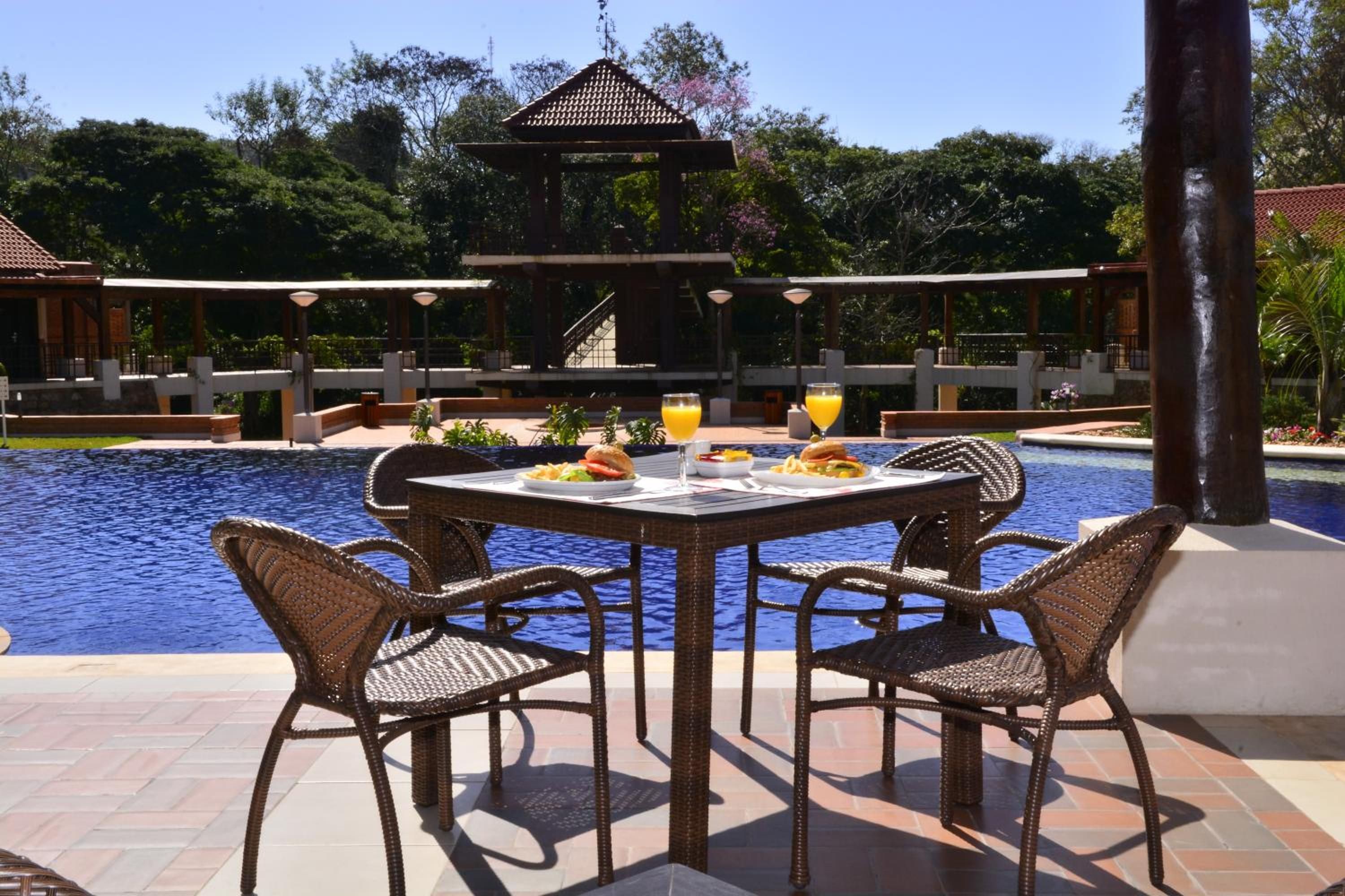Un área de comedor junto a la piscina con sillas de mimbre y una mesa, rodeada por una piscina y un pabellón.
