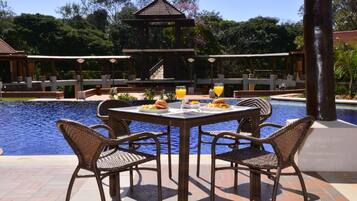 Un área de comedor junto a la piscina con sillas de mimbre y una mesa, rodeada por una piscina y un pabellón.