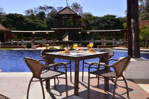 Un área de comedor junto a la piscina con sillas de mimbre y una mesa, rodeada por una piscina y un pabellón.