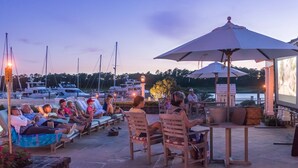 Ein Yachthafen mit Menschen, die auf Liegestühlen entspannen, einem Tisch mit Sonnenschirm, Booten im Hintergrund und einem Bildschirm, der eine Landschaft zeigt.