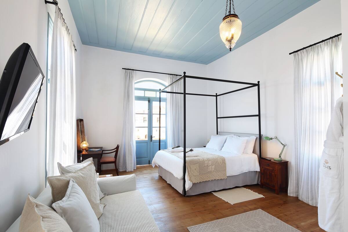 A four-poster bed, a desk with a lamp, a chair, and a large mirror in a bedroom with wooden flooring and a blue ceiling.