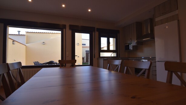 Een ruime eethoek met een lange houten tafel en stoelen, een keuken met moderne apparatuur, en grote ramen met uitzicht op buiten.