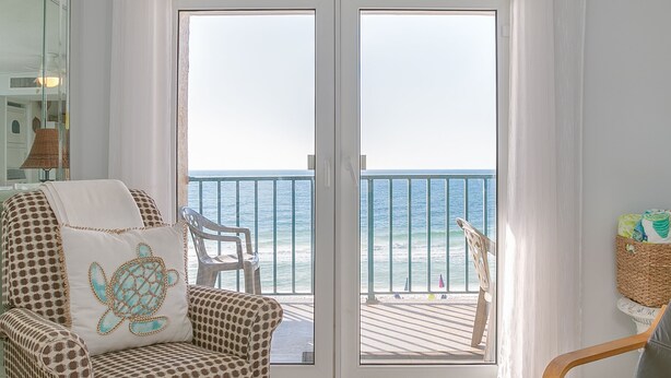 Une pièce dotée d’une grande porte-fenêtre coulissante qui donne sur un balcon offrant une vue sur l’océan. On y trouve un fauteuil à motifs avec un coussin en forme de tortue, une chaise en bois et un panier en osier sur le balcon.