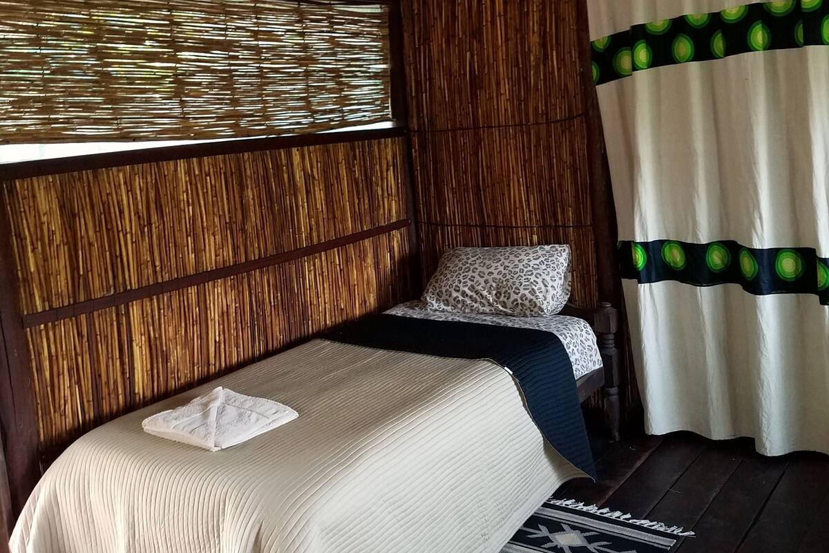 A room with a bed, a window with bamboo blinds, a curtain with a green pattern, and a patterned rug.