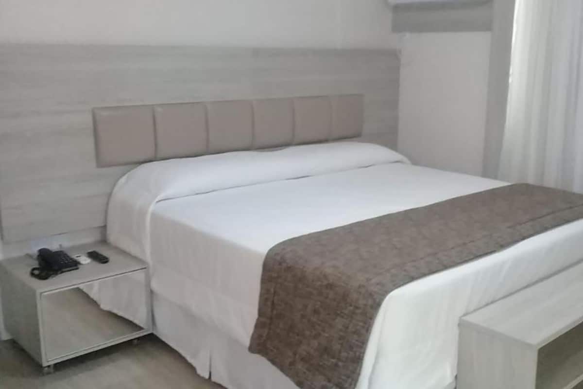 Un lit bien fait, recouvert d’une couvre-lit blanche et marron, une table de chevet avec un téléphone et une télécommande, et une tête de lit en bois clair.