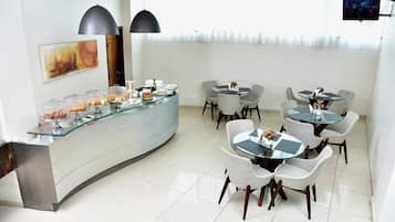 A modern hotel lobby with a curved reception desk, glass display cases with food items, and multiple round tables with chairs.