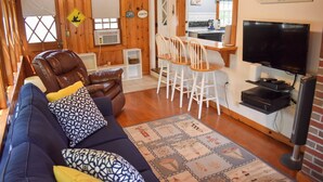 A cozy living room with a leather armchair, a blue sofa with patterned cushions, a coffee table, and a TV. There's a kitchen area with a sign reading "Beach House" and a dining set. - Two bedroom home located less than 2/10's of a mile to shared/private beach (Dennis Port)