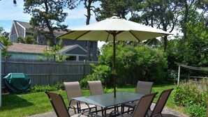 An outdoor patio with a table and chairs, a large umbrella, and a wooden fence in the background. - Two bedroom home located less than 2/10's of a mile to shared/private beach (Dennis Port)