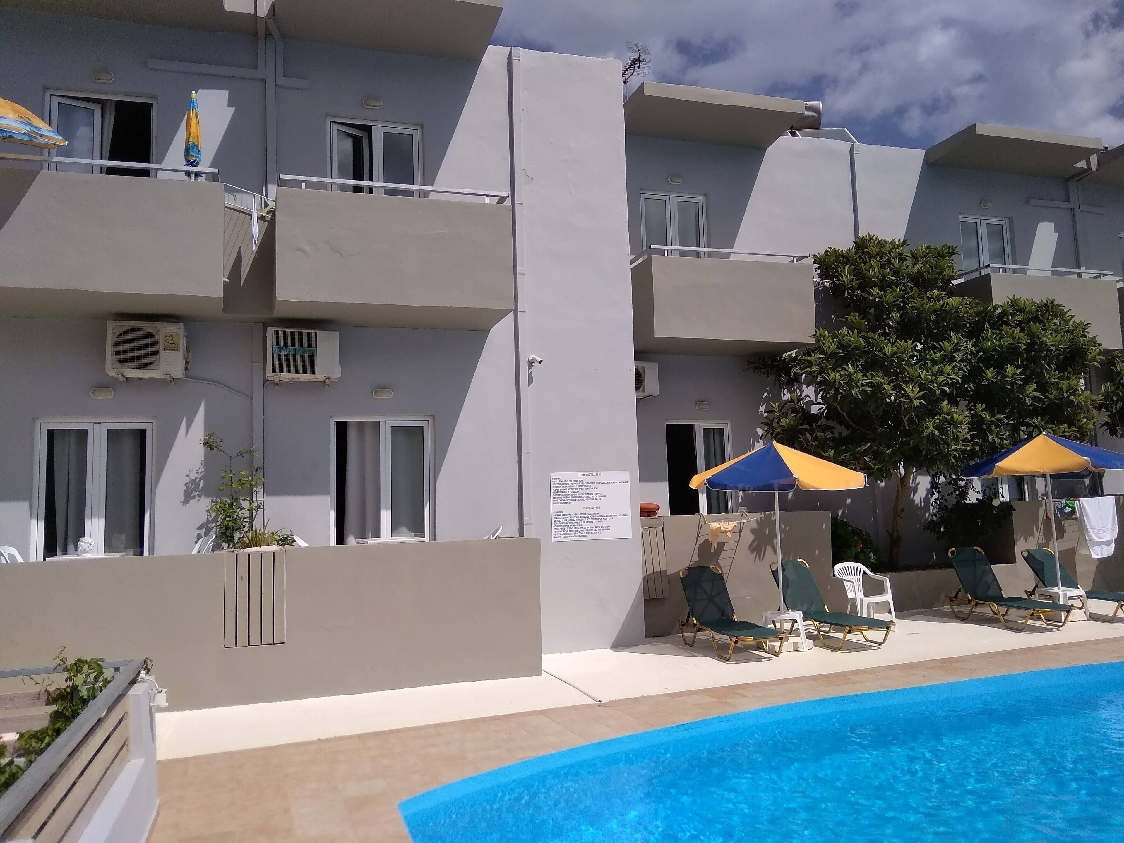 Un edificio moderno con balcones, unidades de aire acondicionado y un área de piscina con sillones de descanso y sombrillas.