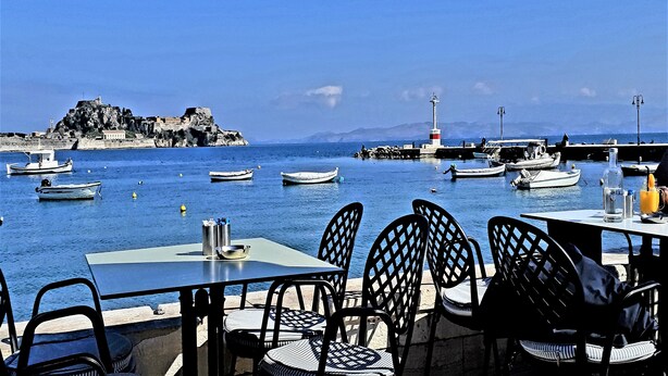 A seaside restaurant with outdoor seating overlooking the water, featuring tables and chairs, a lighthouse, and boats. - Corfu-town Apartment best location/2BR/lift/removed/smart<br>TV+Netflix.<br>  (Kerkira)
