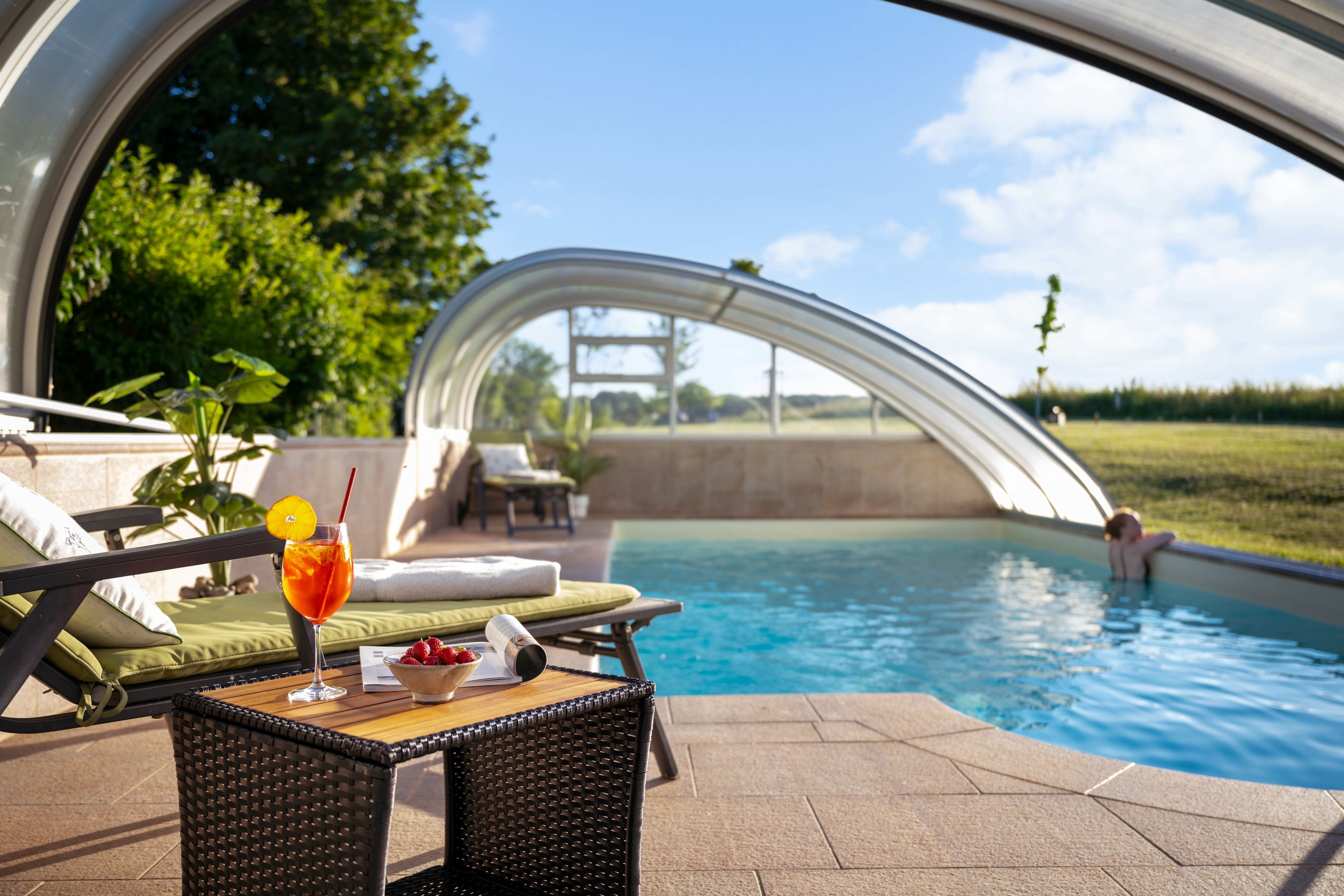 Un espace aménagé au bord de la piscine, avec un fauteuil de relaxation protégé par des parois de verre, une table avec des boissons et des fruits, et une vue sur la piscine et le paysage environnant.