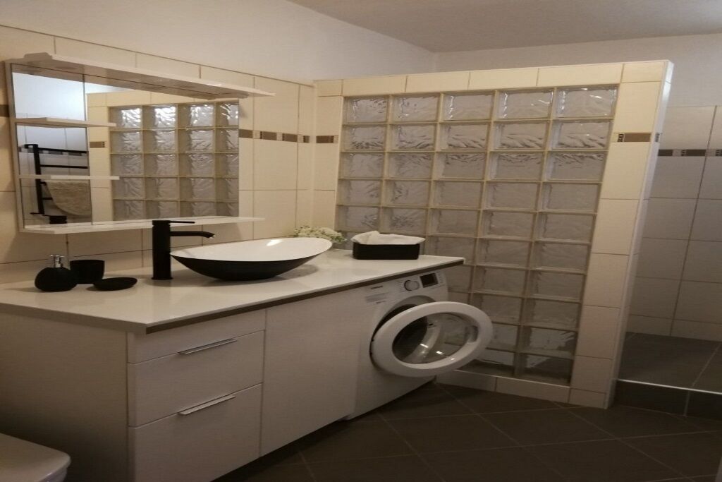 Une salle de bain moderne avec un grand miroir, un lavabo doté d’un robinet noir, un lave-linge et une cabine de douche carrelée.