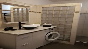 Une salle de bain moderne avec un grand miroir, un lavabo doté d’un robinet noir, un lave-linge et une cabine de douche carrelée.