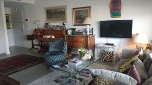 A living room with a sofa, armchair, coffee table, and a TV. There is a bookshelf, a desk, and a colorful artwork on the wall. - Beautiful 1 Bedroom in New GINDI Complex (Tel Aviv-Yafo)