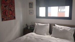 A bedroom with a large bed, a window with blinds, a nightstand with books, and a wall with a framed picture and a painting. - Beautiful 1 Bedroom in New GINDI Complex (Tel Aviv-Yafo)