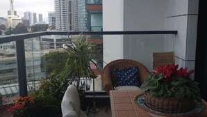 A balcony with a view of a cityscape, a wicker chair, a table with a basket of flowers, and a potted plant. - Beautiful 1 Bedroom in New GINDI Complex (Tel Aviv-Yafo)