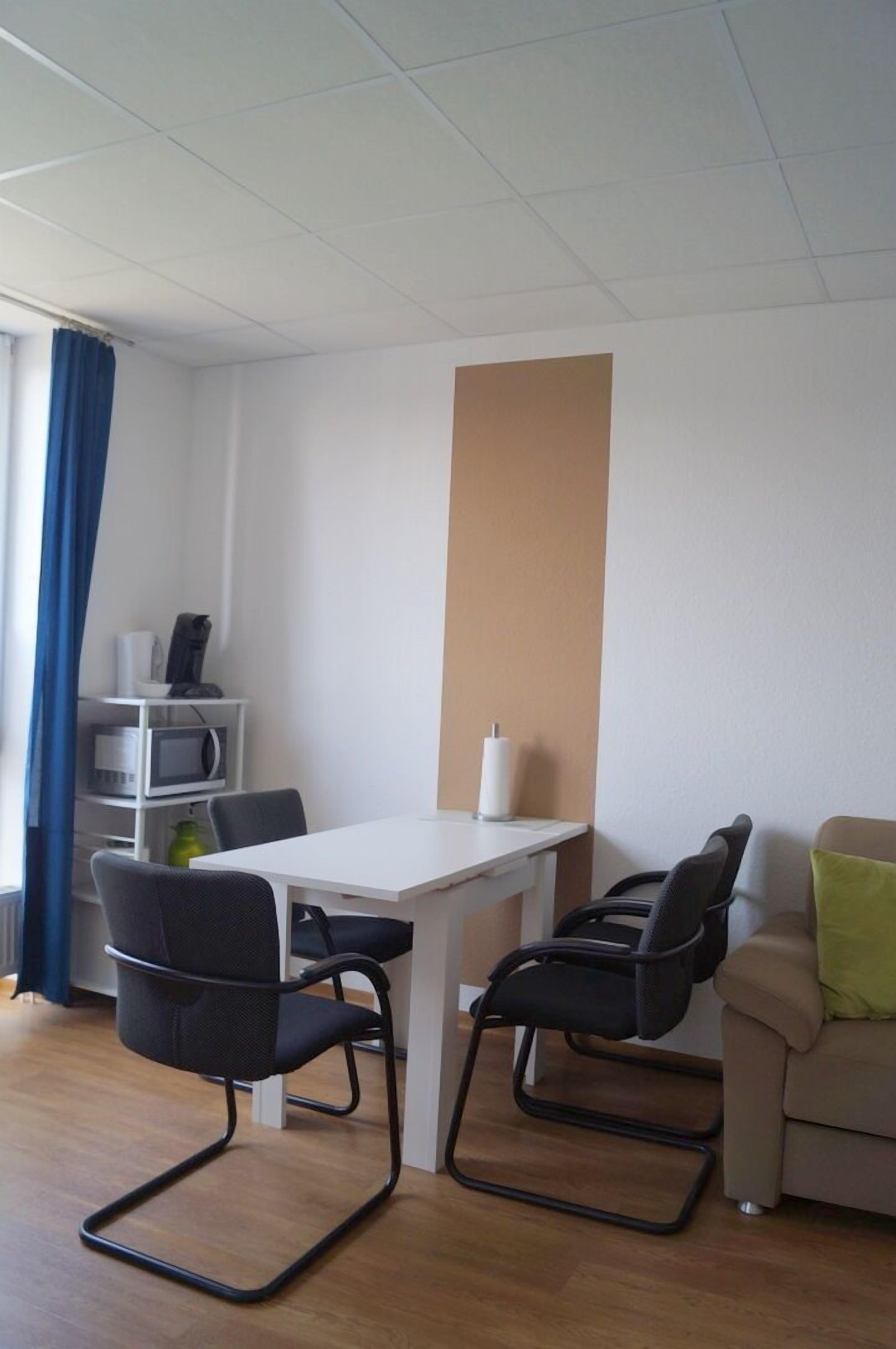 Quarto moderno com uma mesa branca, duas cadeiras pretas, um sofá bege e uma cortina azul.