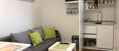 Un espacio habitable compacto con un sofá gris, una pequeña mesa blanca, una kitchenette con microondas y un cuadro en la pared.