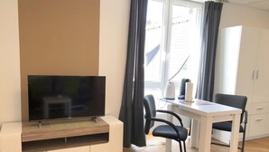 Een moderne woonkamer met een flatscreen televisie op een houten standaard, een eettafel met twee stoelen, een witte kast en een raam met gordijnen.