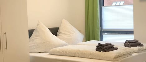 Una cama bien hecha con almohadas y toallas dobladas, una ventana con persianas y una cortina verde.