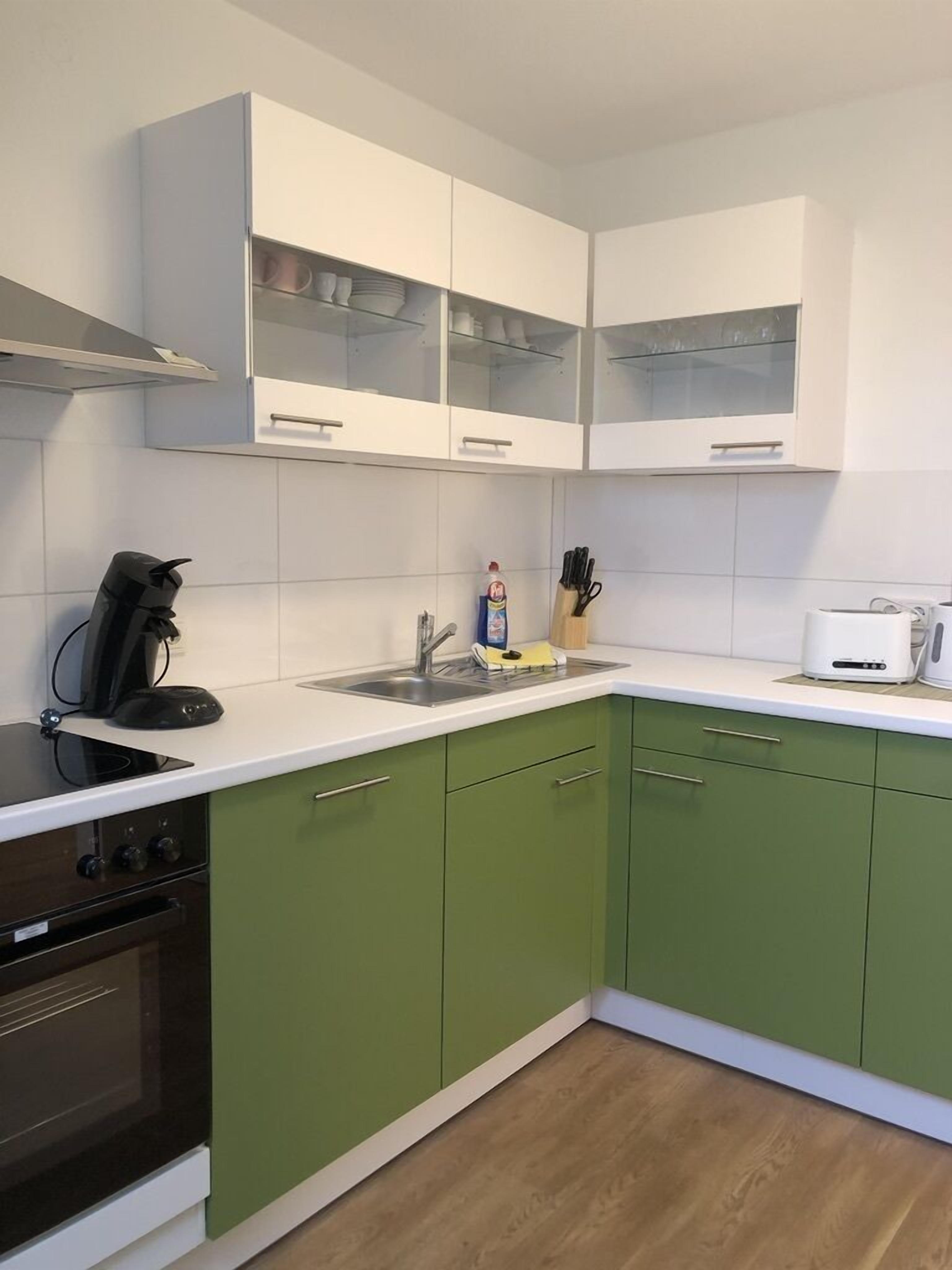 Een keuken met groene kastjes, witte aanrechtbladen, een zwarte kookplaat, een koffiezetapparaat, een broodrooster en een bovenkast met glazen deuren.