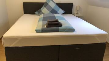 Una cama con cabecera oscura, almohada a cuadros y una toalla doblada sobre un colchón blanco.
