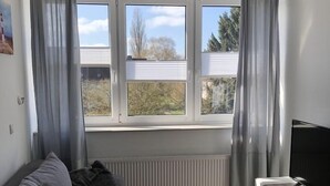 Een moderne woonkamer met een grijze bank, een klein wit tafeltje, een televisie op een houten dressoir en een raam met gordijnen.