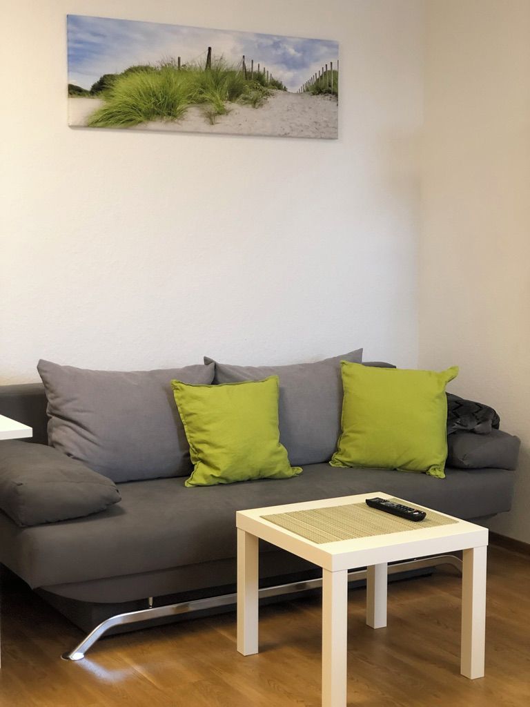 Ein Wohnzimmer mit grauem Sofa, zwei grünen Kissen, einem weißen Couchtisch und einem Strandbild an der Wand.