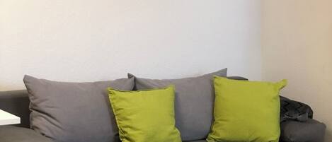 Ein Wohnzimmer mit grauem Sofa, zwei grünen Kissen, einem weißen Couchtisch und einem Strandbild an der Wand.
