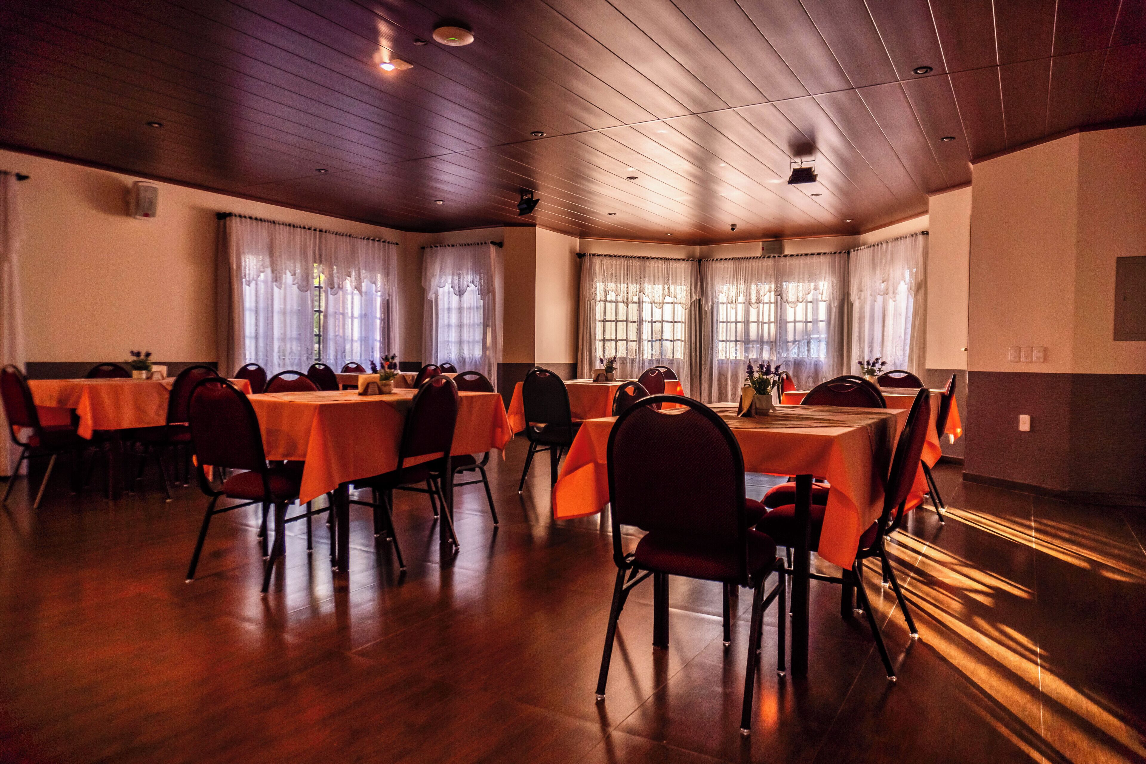 A dining area with multiple tables and chairs, wooden flooring, and large windows with curtains.
