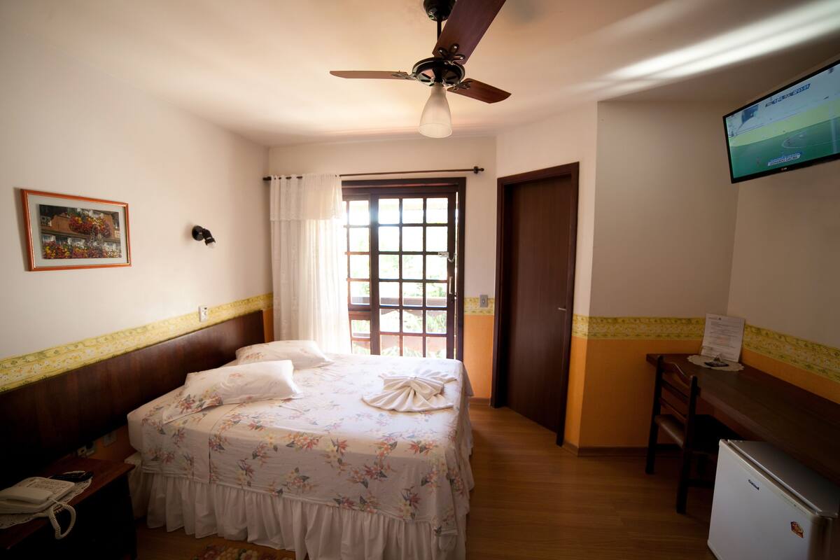 A bedroom with a bed, a desk, a refrigerator, a ceiling fan, a window with curtains, and a television on the wall.