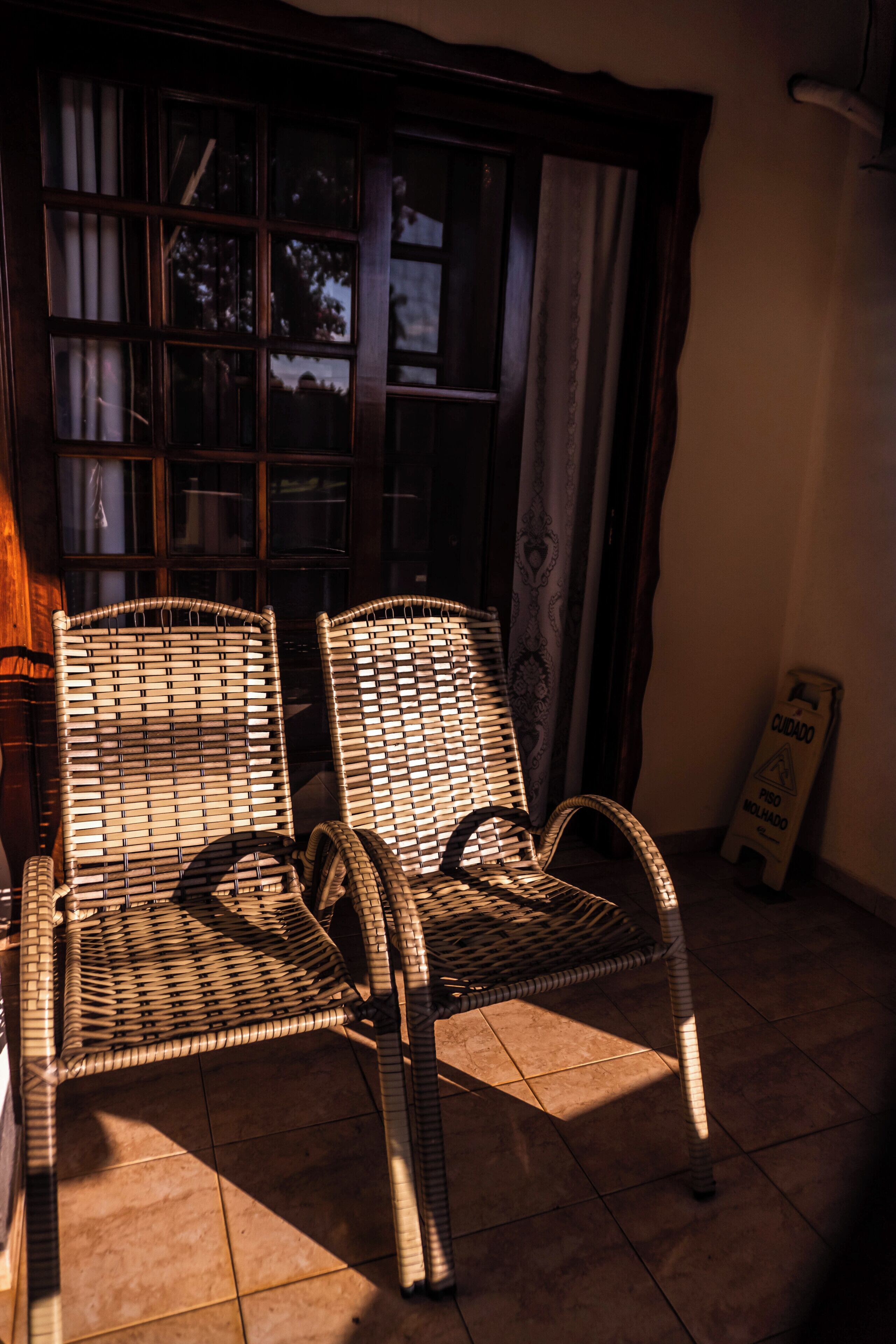 Two wicker chairs near a window with curtains, a tiled floor, and a sign on the wall.