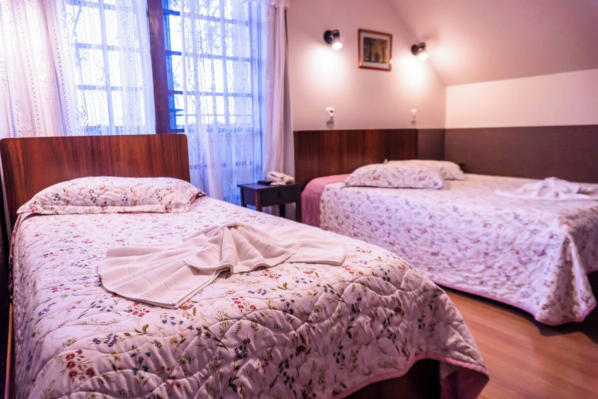 A hotel room with two beds, a wooden headboard, a bedside table with a phone, and a window with sheer curtains.