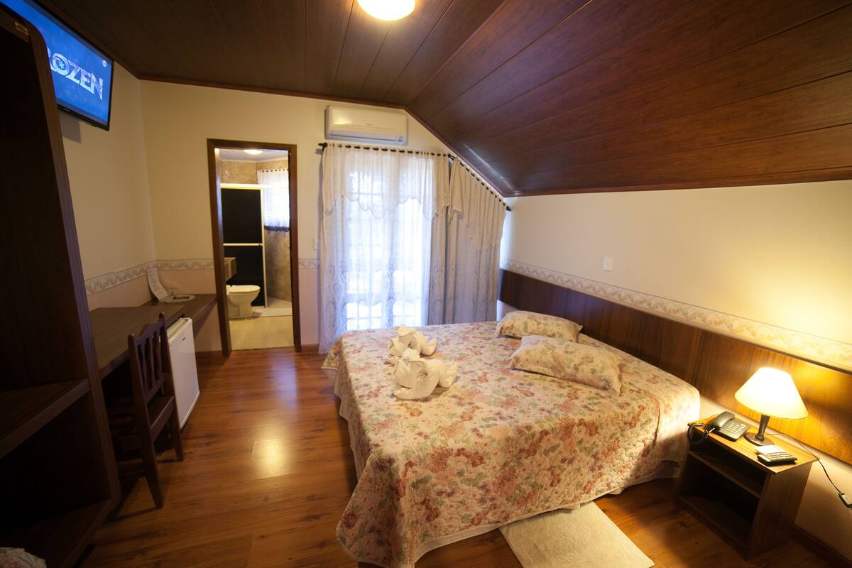 A hotel room with a wooden ceiling, a bed with floral bedding, a bedside table with a lamp, a desk with a chair, and an air conditioning unit.