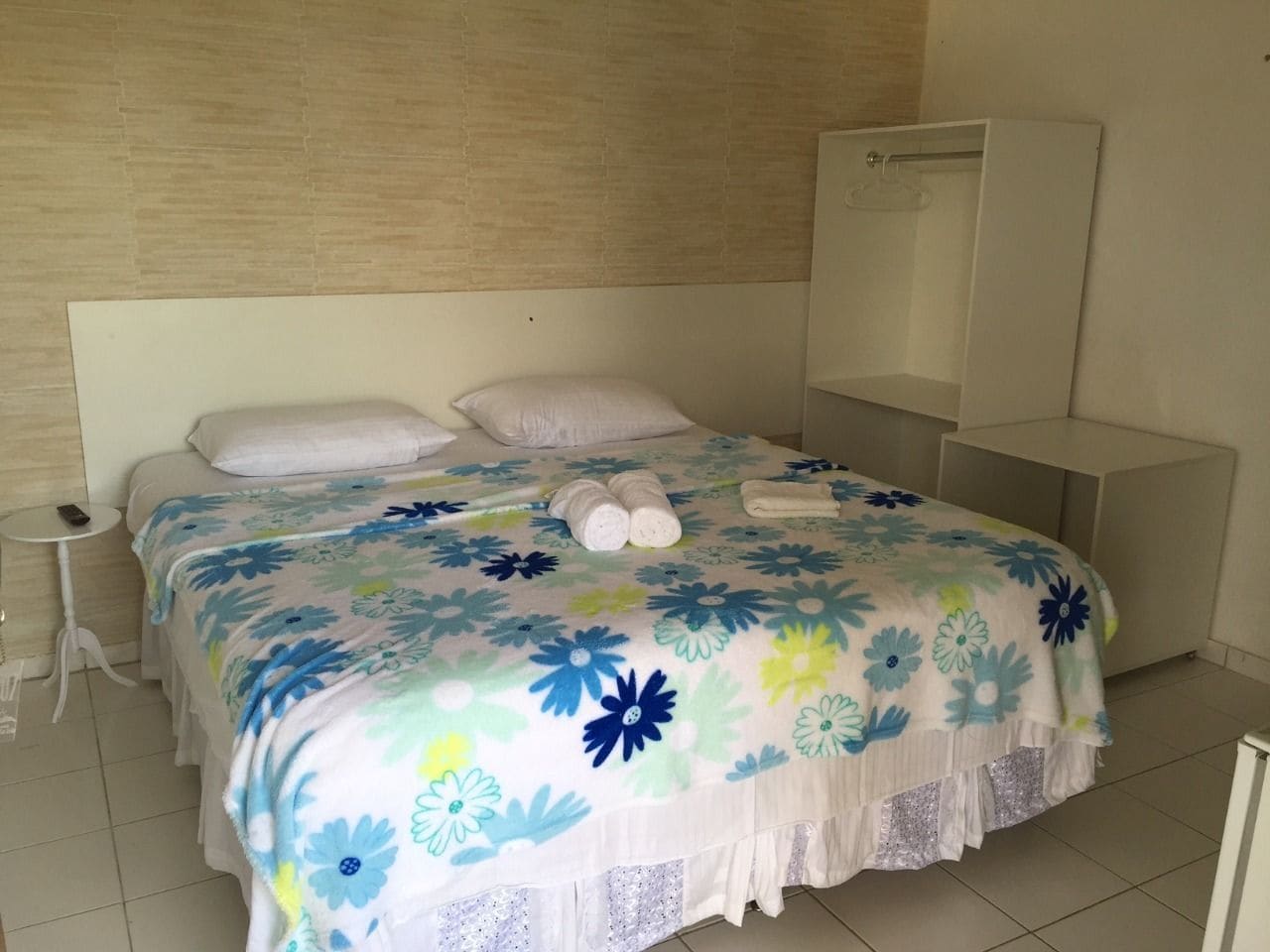 A neatly made bed with a floral bedspread, a bedside table with a remote control, and a wardrobe in a hotel room.