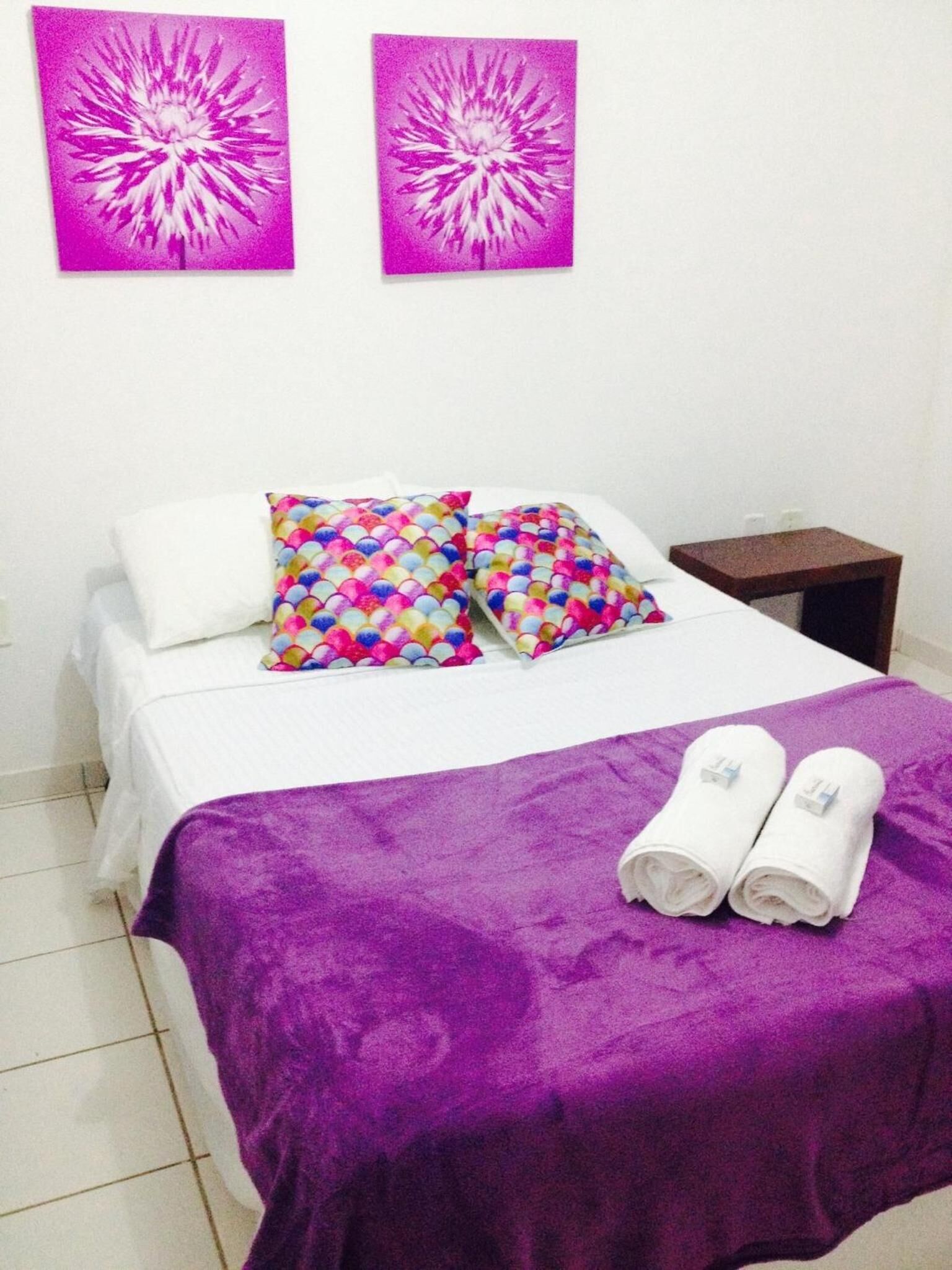 A neatly made bed with white linens, a purple blanket, and colorful pillows. There are two rolled towels on the bed. The room has white walls and a small wooden nightstand beside the bed.