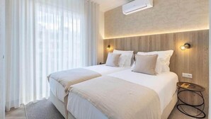 Hotellihuone, jossa on kaksi sänkyä, suuri ikkuna läpikuultavilla verhoilla, ilmastointilaite ja pieni yöpöytä, jossa on lamppu.