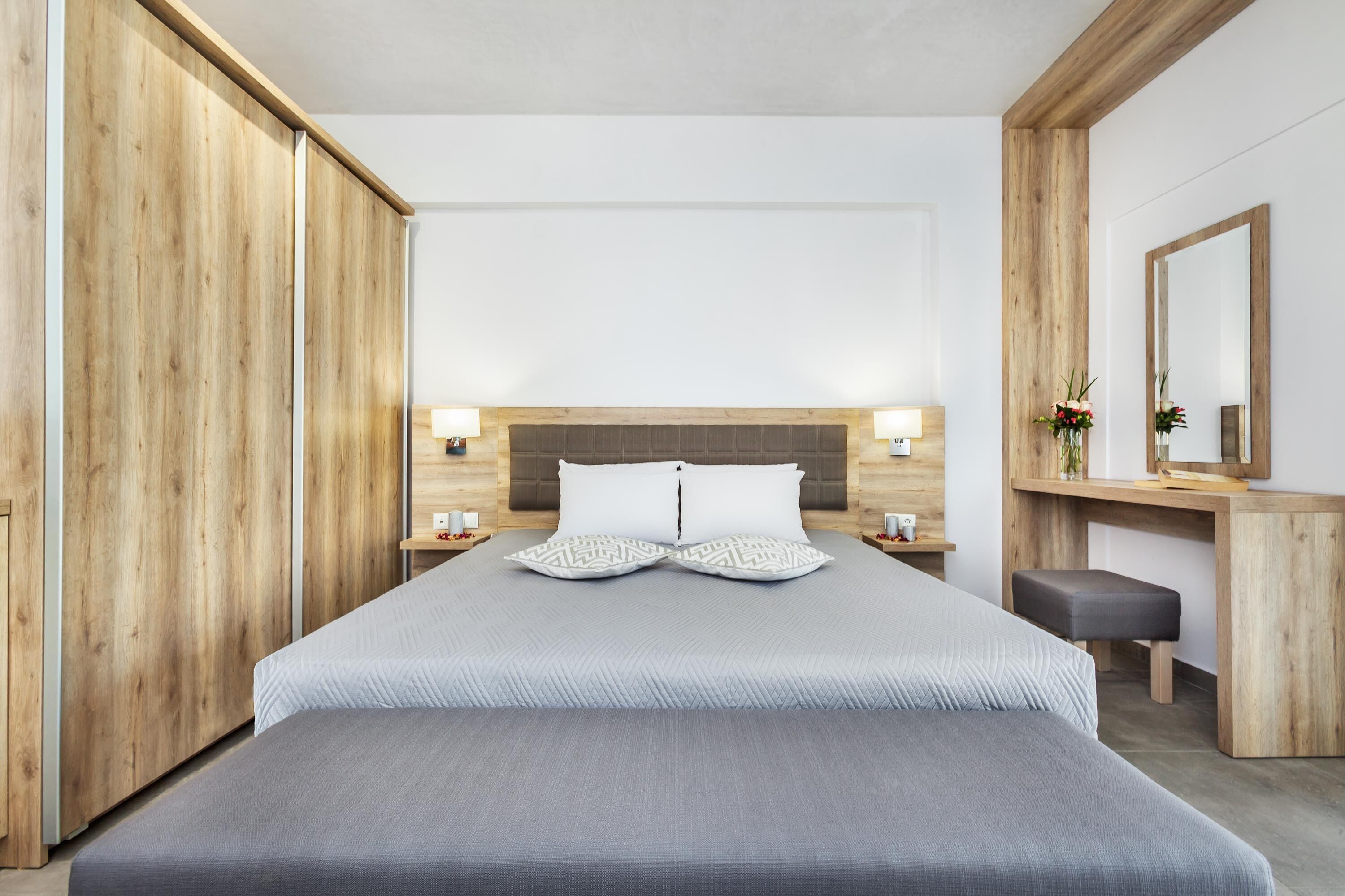 A modern hotel room with a large bed, wooden wardrobe, bedside tables with lamps, a desk with a mirror, and a bench.