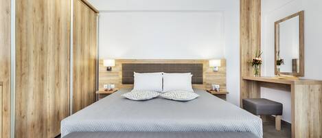 A modern hotel room with a large bed, wooden wardrobe, bedside tables with lamps, a desk with a mirror, and a bench.