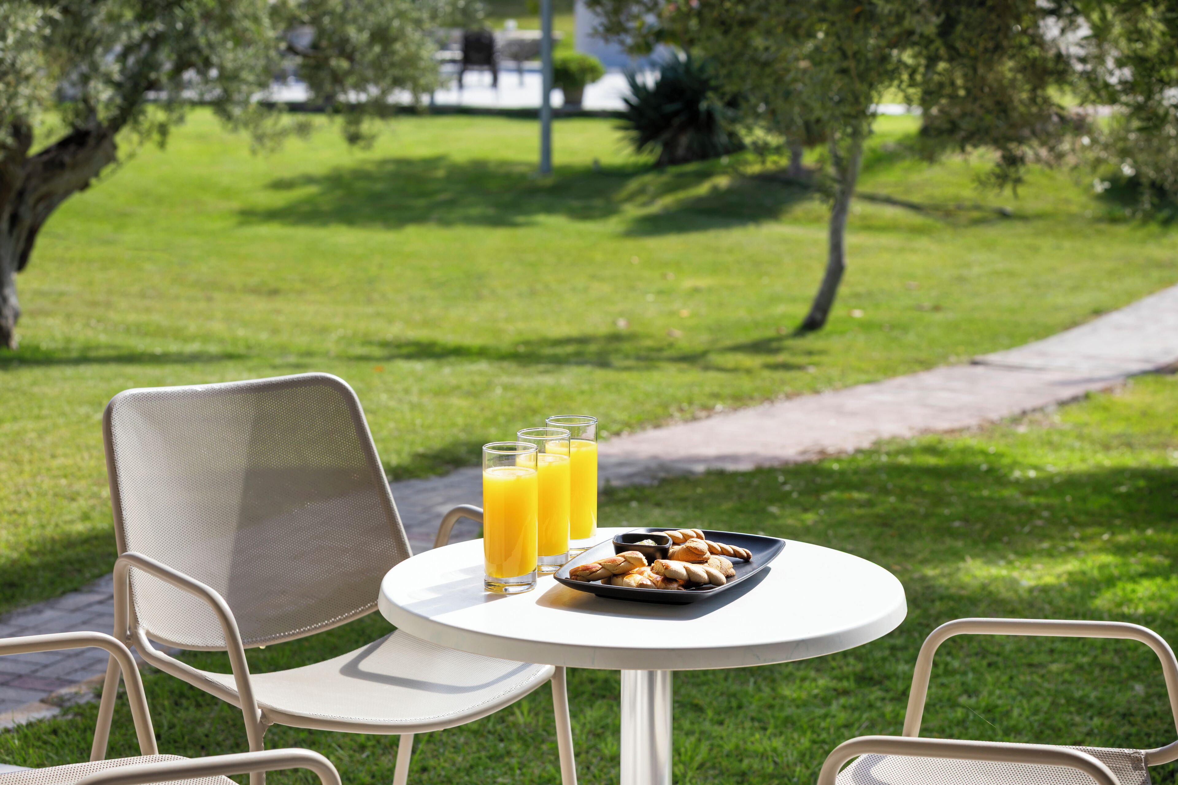 A table with chairs and a tray of pastries and orange juice in a grassy outdoor setting.