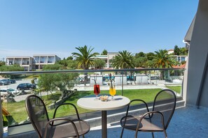 A balcony with two wicker chairs, a small round table, and two drinks. The view includes a parking area, a swimming pool, and buildings with palm trees. - Iris Hotel (Kassandra)