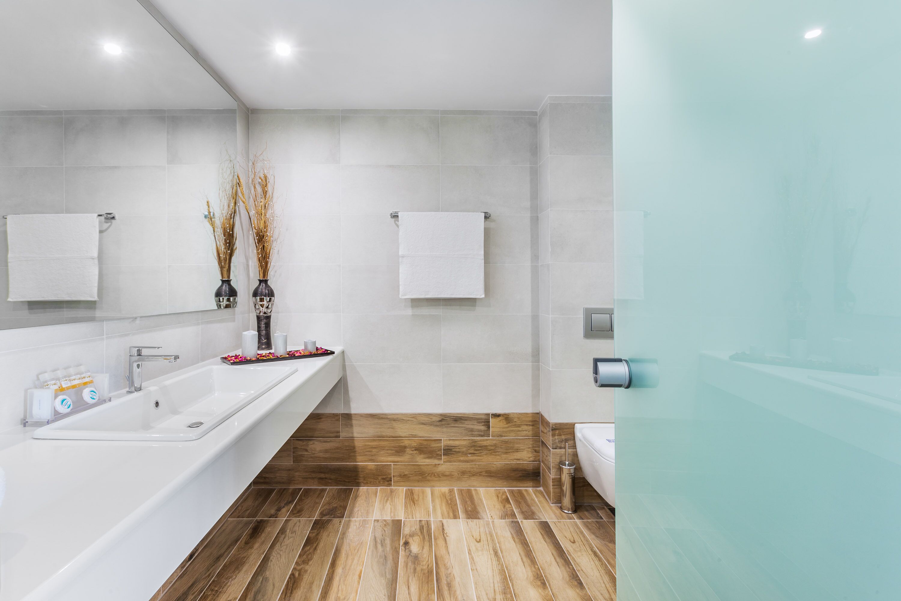 A modern bathroom with a long white sink, two white towels, and a glass shower enclosure.