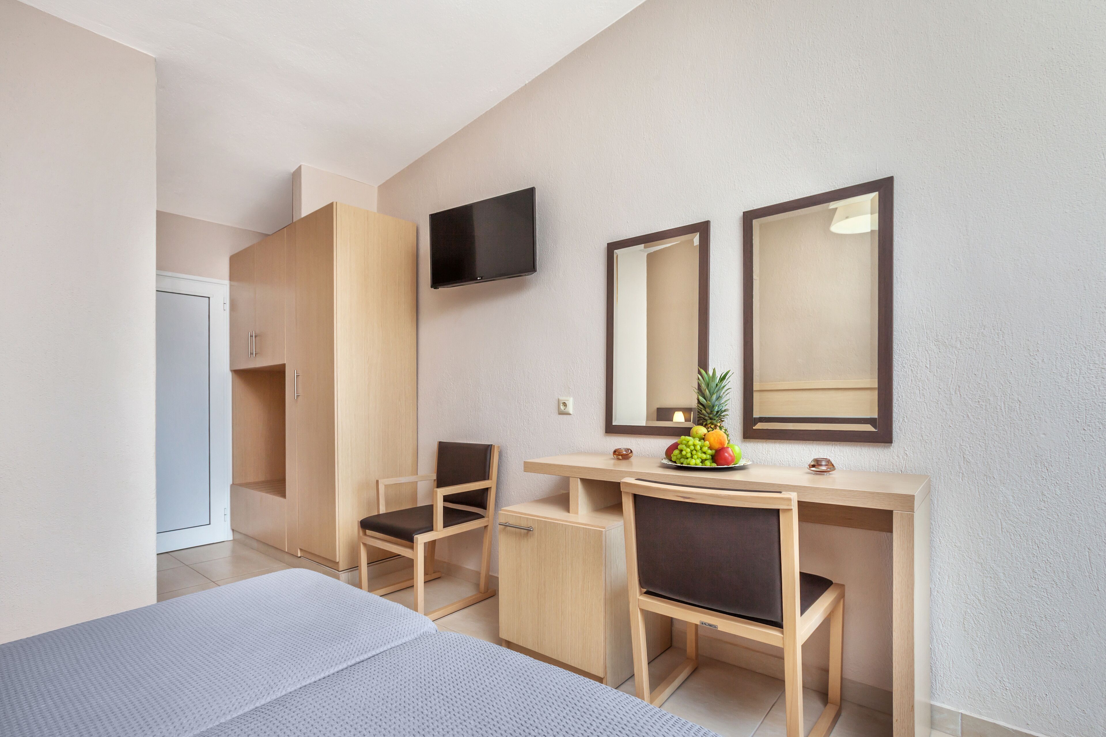 A hotel room with a bed, a desk with two chairs, a television, and a fruit bowl on the desk.