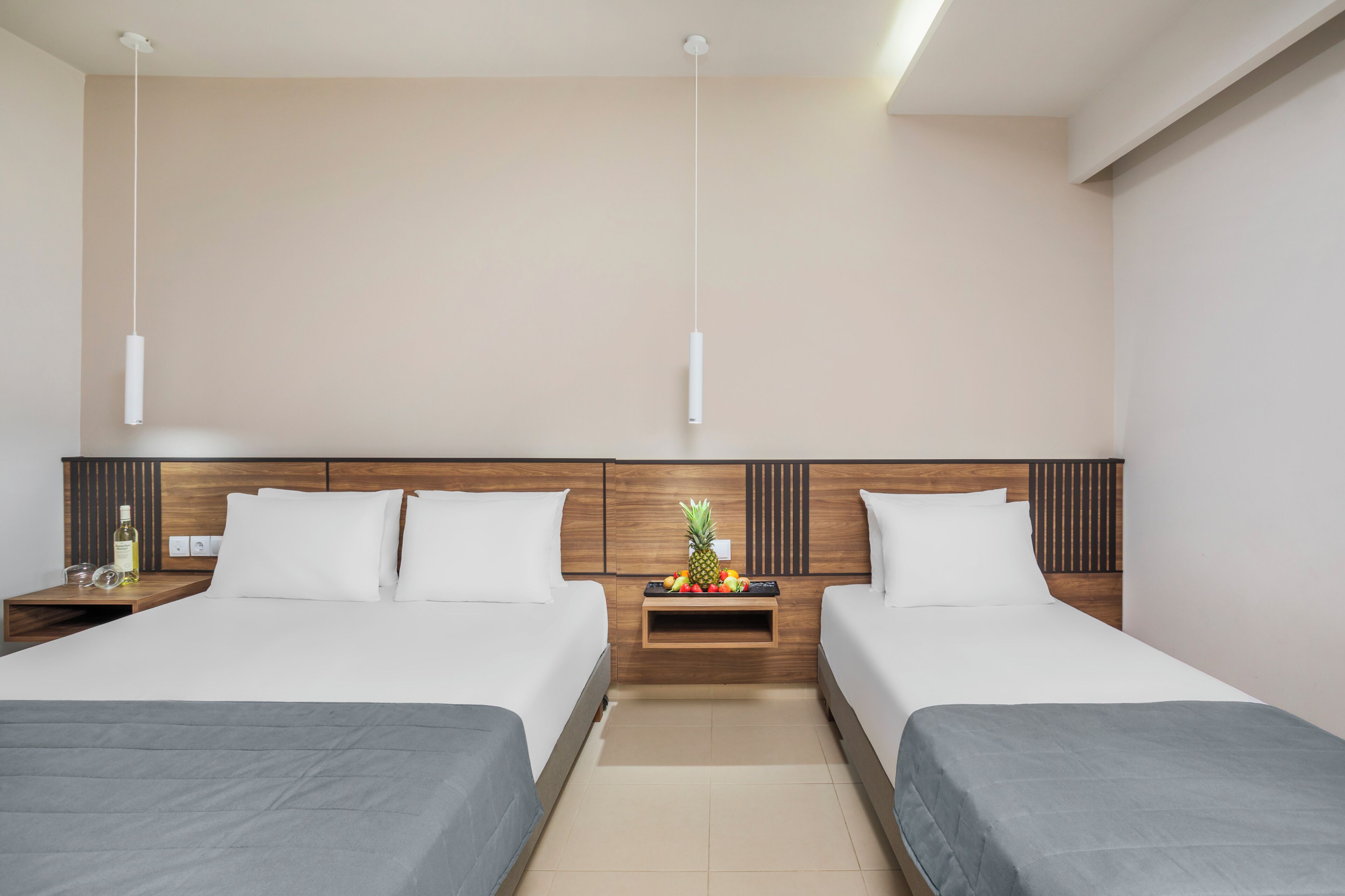 A hotel room with two beds, a wooden headboard, a small table with fruit, and a nightstand with a bottle and glasses.