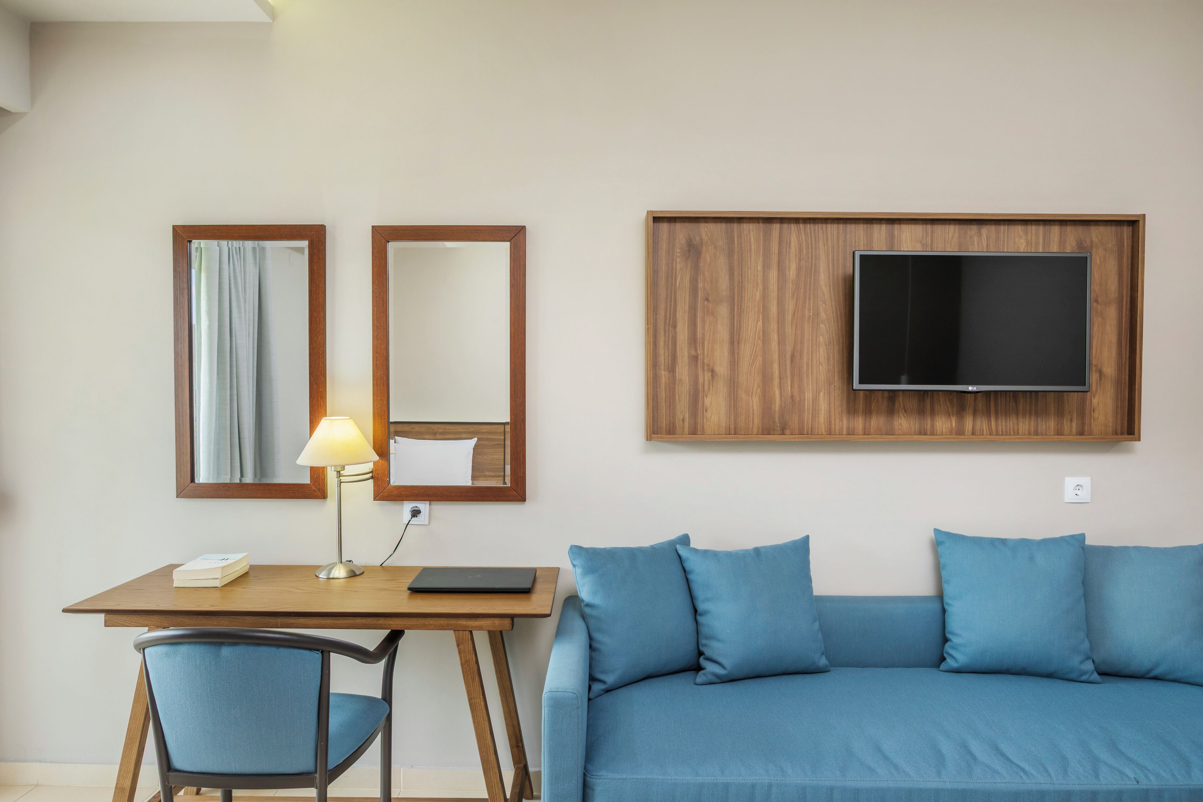 Una habitación con un escritorio de madera, un sofá azul, dos espejos, una lámpara y un televisor de pantalla plana montado en la pared.