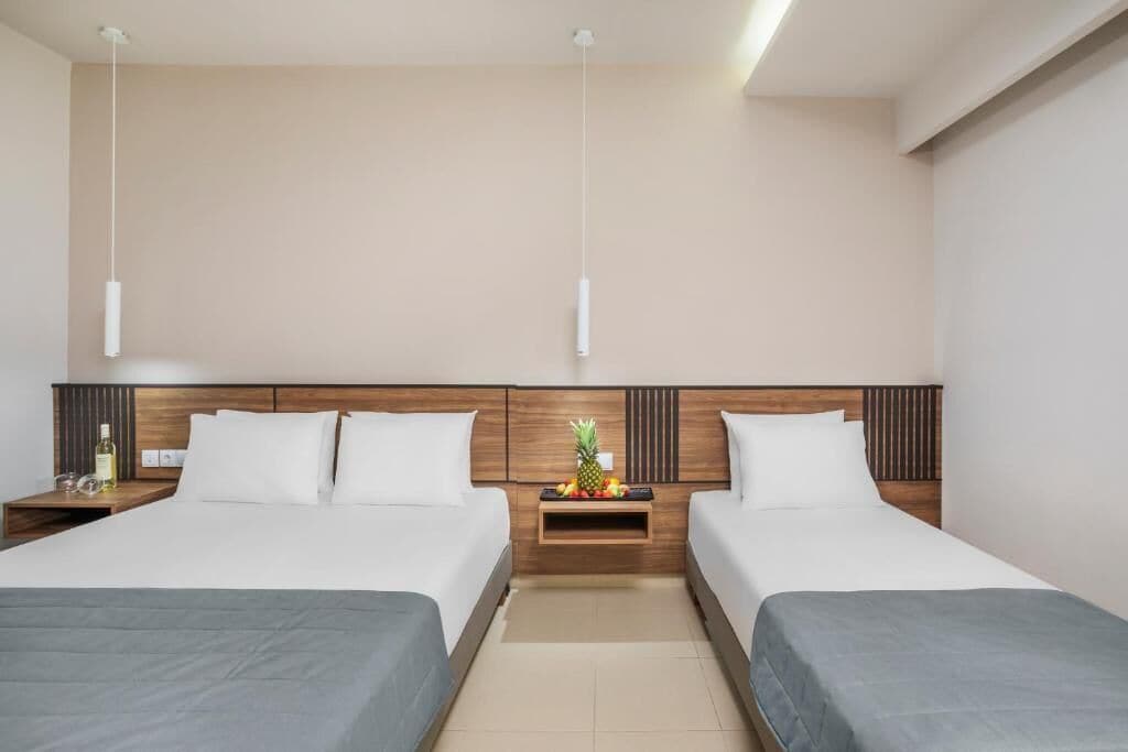 A hotel room with two beds, wooden headboards, a nightstand with a fruit bowl, and pendant lights.