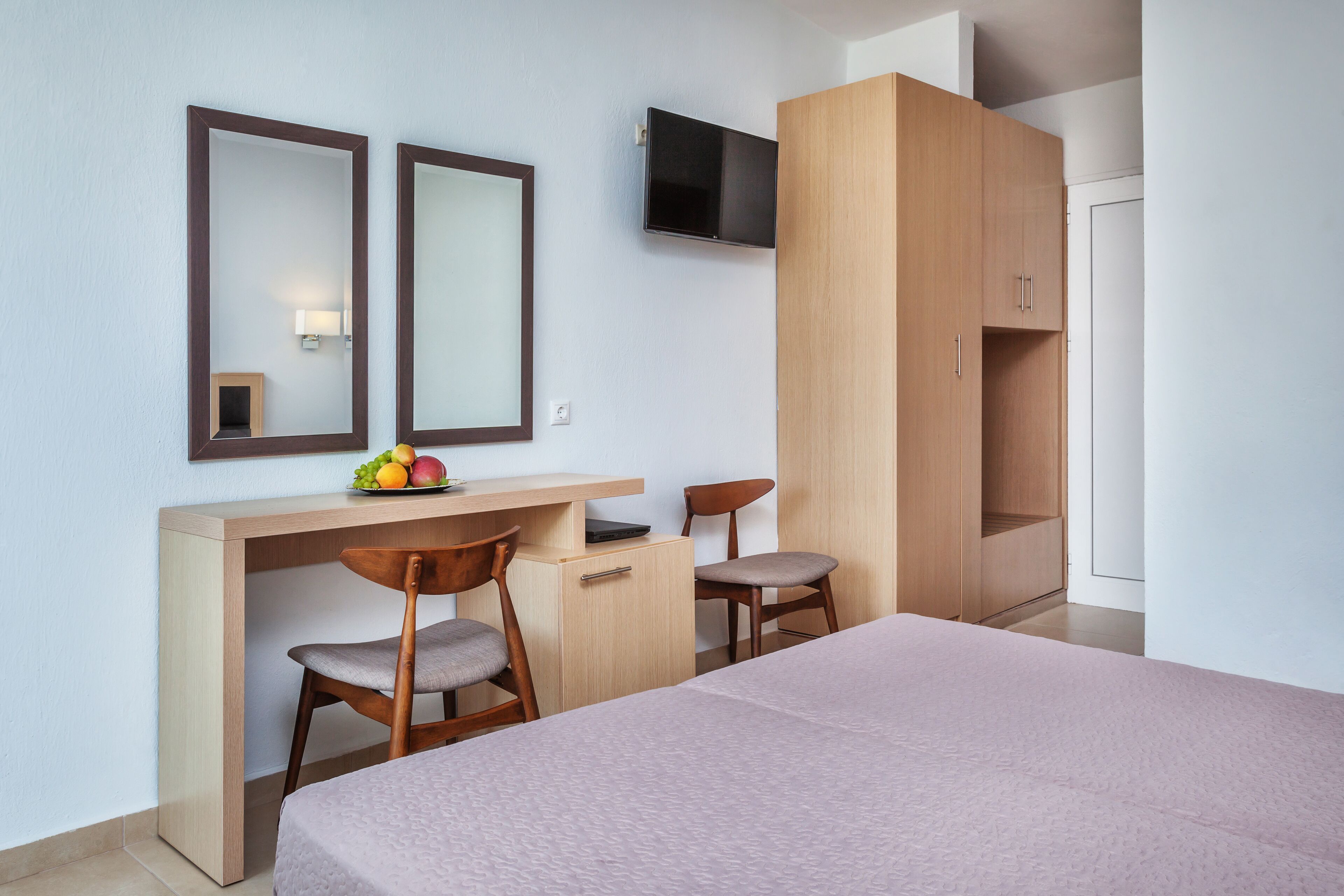 Una habitación de hotel con cama, escritorio con dos sillas, televisor montado en la pared y armario.